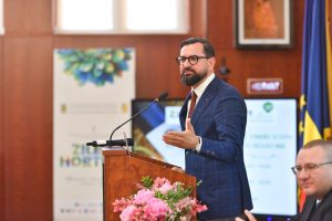 Adrian Chesnoiu vorbind la o tribună din lemn decorată cu flori roz, în timpul unui eveniment oficial. În fundal se observă un banner cu sigla Zilele Horticulturii și steagul României.