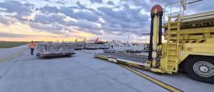 Prim-plan cu o platformă galbenă de ridicare a încărcăturii cargo (loader) pe o pistă de aeroport betonată la apus. În fundal se observă palete cu marfă și coada unui avion.