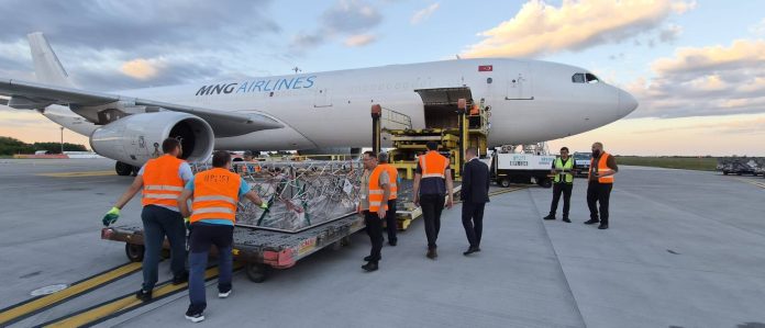 Lucrători la sol în veste portocalii de siguranță ghidând o paletă de marfă pe o platformă de ridicare galbenă către ușa deschisă a unui avion MNG Airlines pe o pistă la apus.
