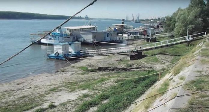 O fotografie panoramică care surprinde malul Dunării în timpul unei perioade de secetă severă. Nivelul scăzut al Dunării a retras apa, expunând o fâșie lată de nisip și vegetație uscată între malul înalt și linia apei. Mai multe pontoane de acostare și structuri plutitoare albastre și albe sunt eșuate pe uscat sau în apă foarte puțin adâncă, cu pasarelele lungi de metal întinse peste zona uscată pentru a ajunge la ele. În fundal, se văd alte ambarcațiuni și macarale portuare pe celălalt mal al fluviului