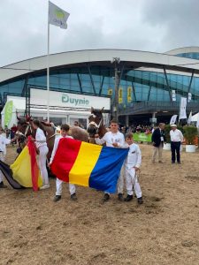 Doi tineri crescători români și un adult arborează steagul României în fața cailor de tracțiune Ardenez la Târgul de la Libramont, Belgia, 2026.